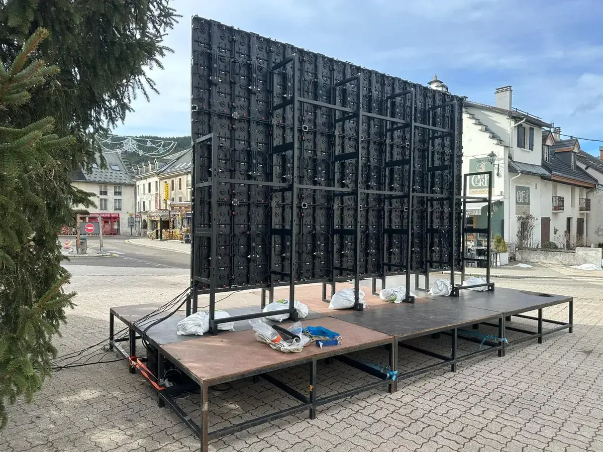 Back of an outdoor LED wall on its steel frame, standing on a stage platform in a village square