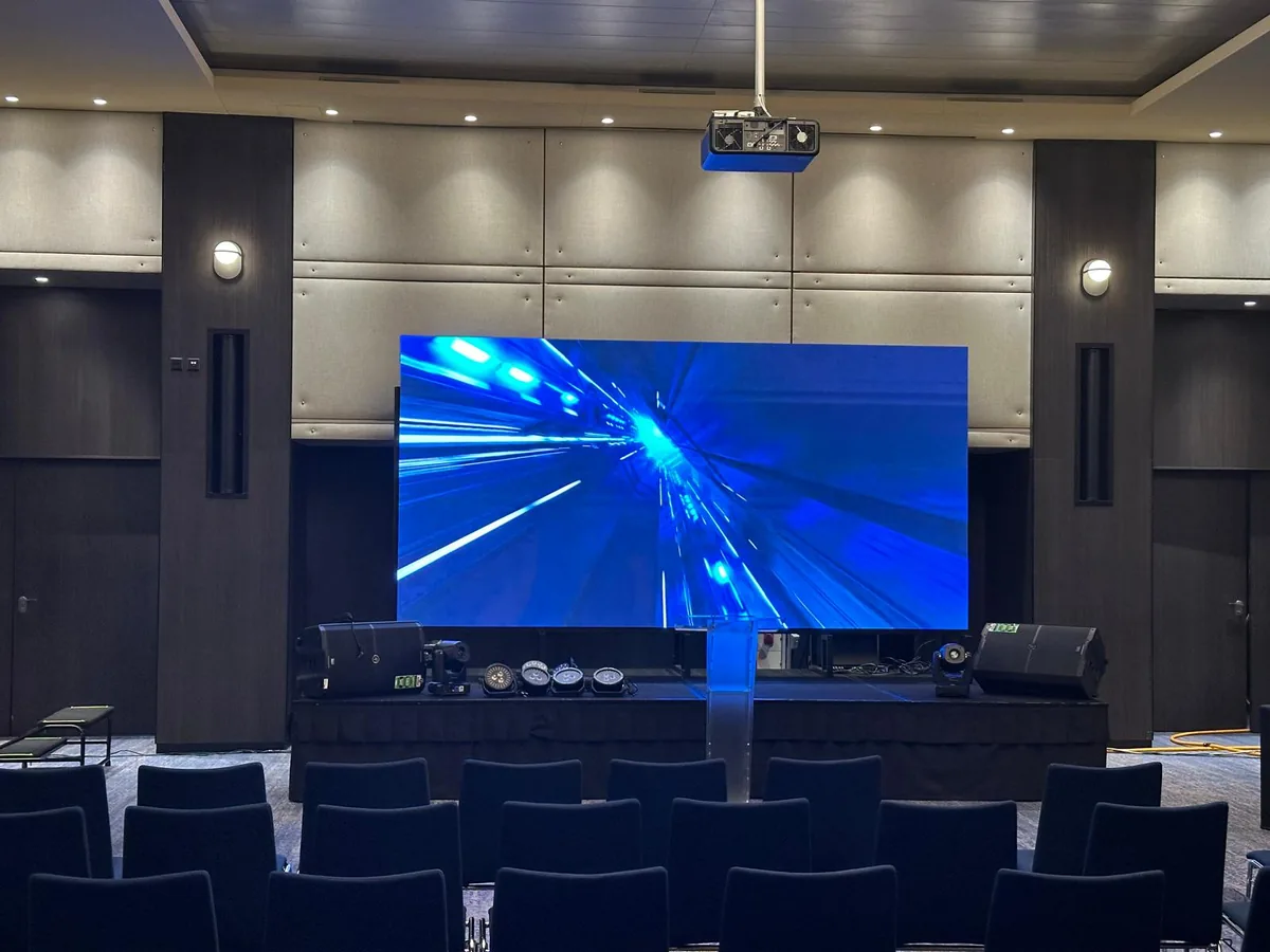 Function room set for a conference: an LED wall lit behind the stage, speakers and small stage lights at its foot, rows of chairs in front