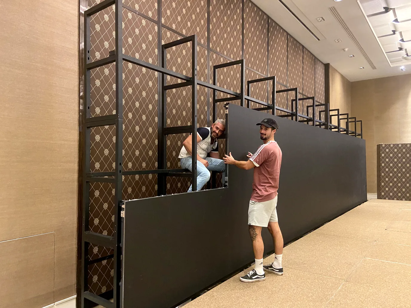 Two technicians fitting black LED panels onto a steel frame in a function room with a patterned wall
