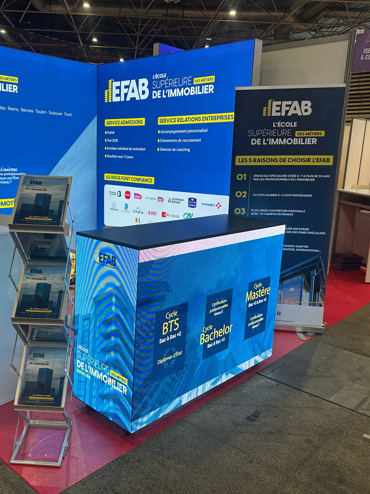 Comptoir d’accueil LED sur un stand de salon, sa façade et son côté montrant les programmes de l’exposant