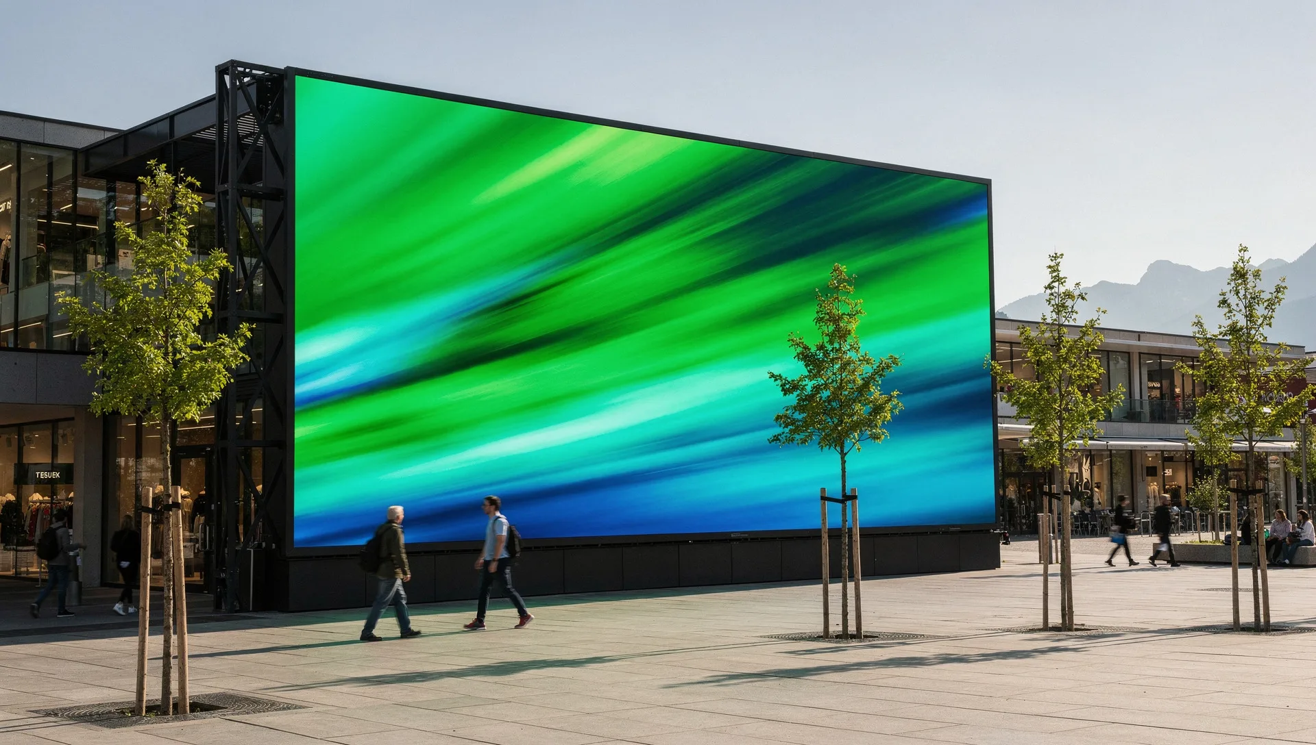Large outdoor LED screen on the side of a building above a paved forecourt with young trees, mountains behind