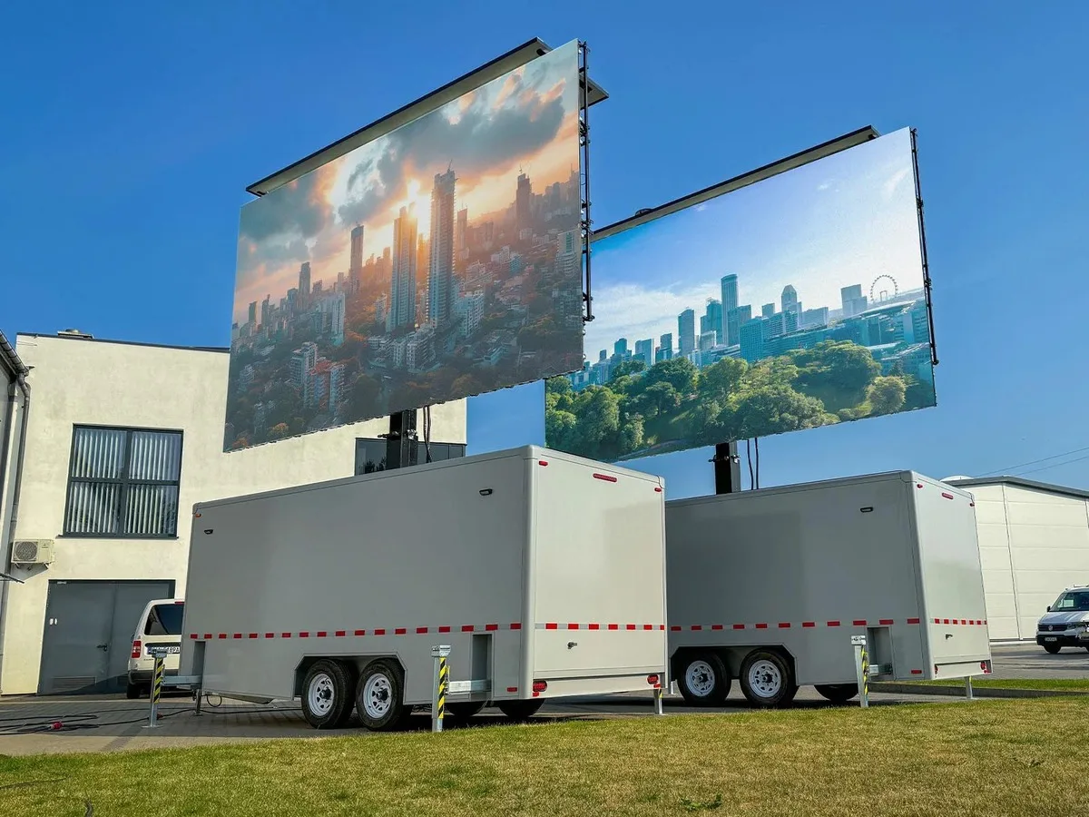 Two LED trailers side by side beside a building, their screens raised and showing city skylines
