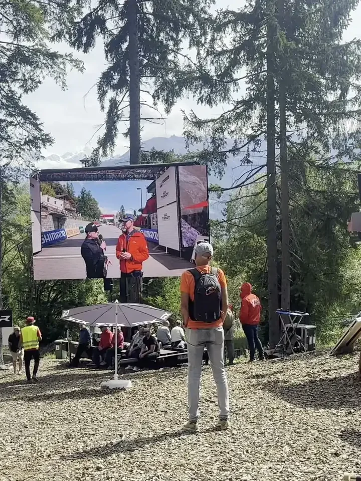 LED trailer screen raised above deckchairs at an outdoor sports event, a spectator walking past