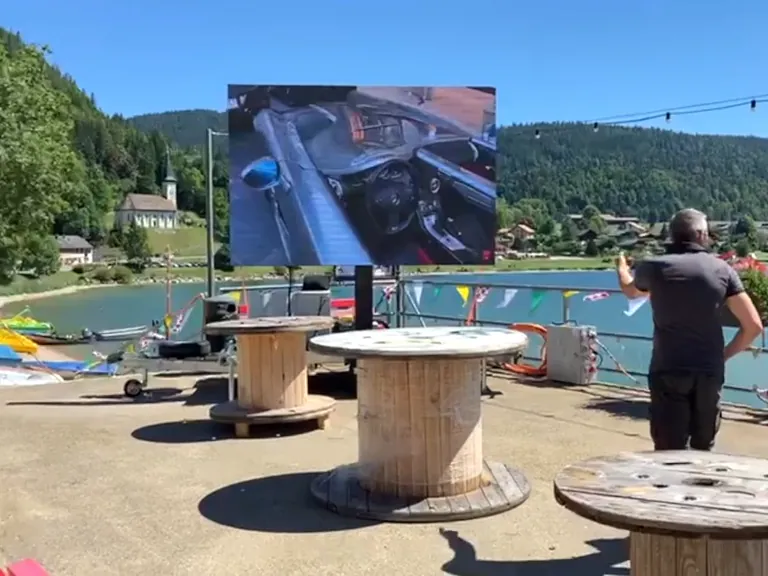 LED trailer screen facing picnic tables by a lake, people watching