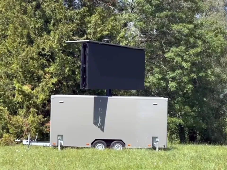 LED trailer in a meadow with its screen raised on its mast, trees behind