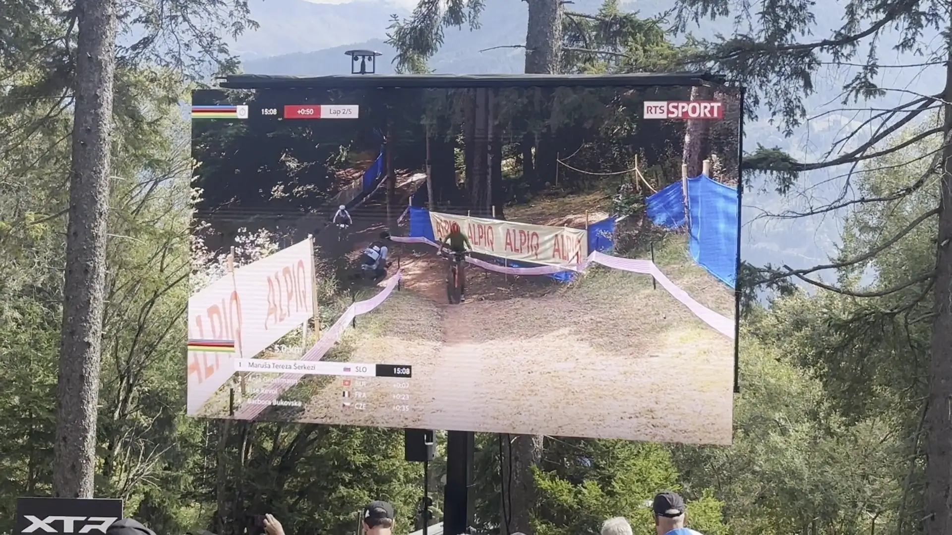 LED trailer screen showing a mountain-bike race to spectators at the edge of a forest