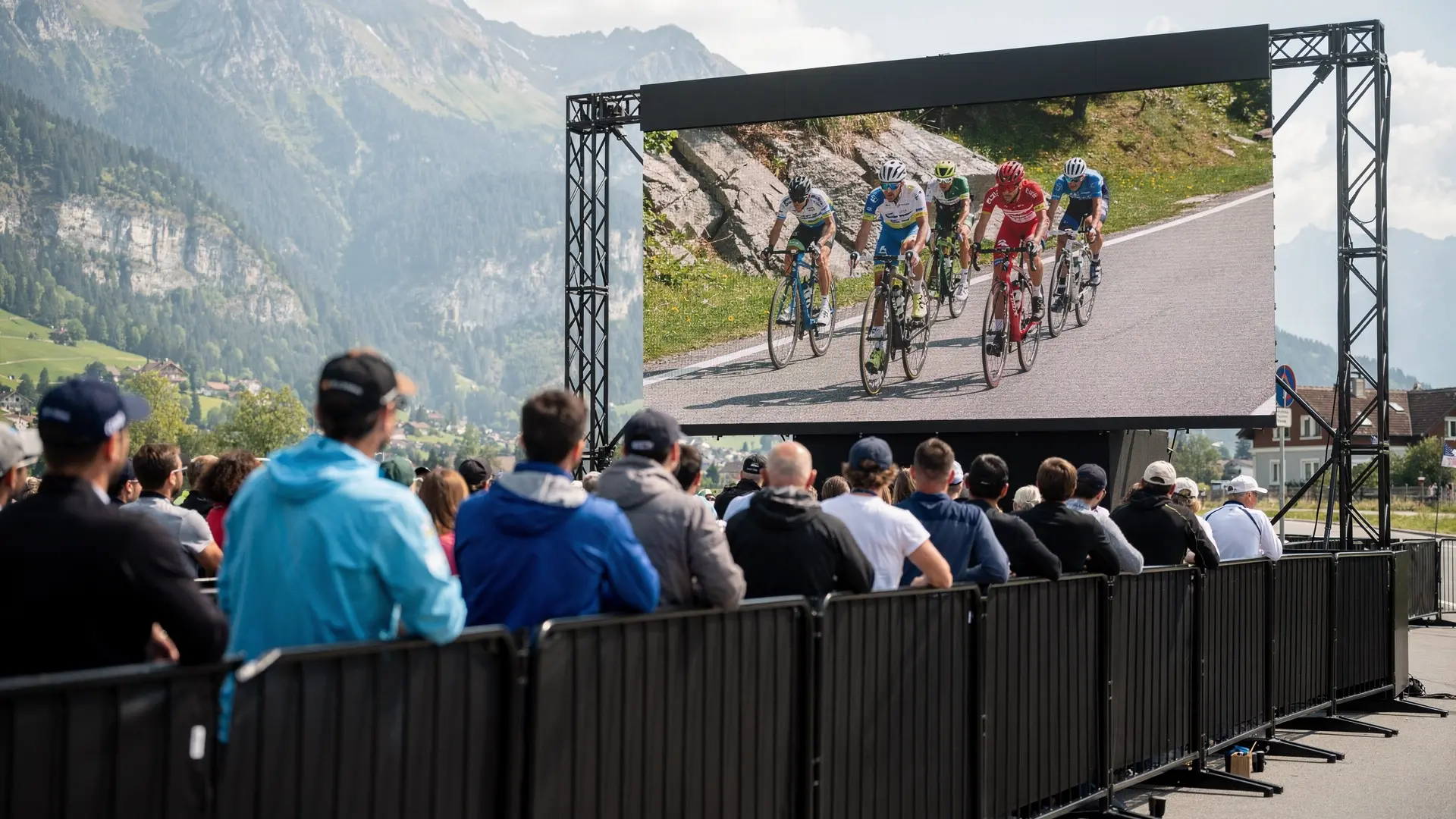 Outdoor LED screen on a truss frame in front of a crowd behind barriers, a cycling race on screen, mountains behind