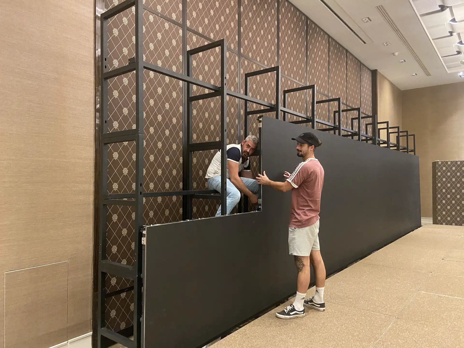 Two technicians fitting black LED panels onto the steel frame of an LED wall, in front of a patterned wall
