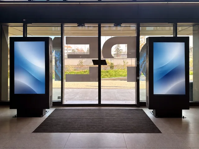 Two digital totems standing either side of a glass entrance door, a doormat between them